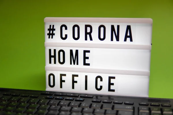 a light box with the inscription: #CORONA HOME OFFICE is behind a black computer keyboard against a green background