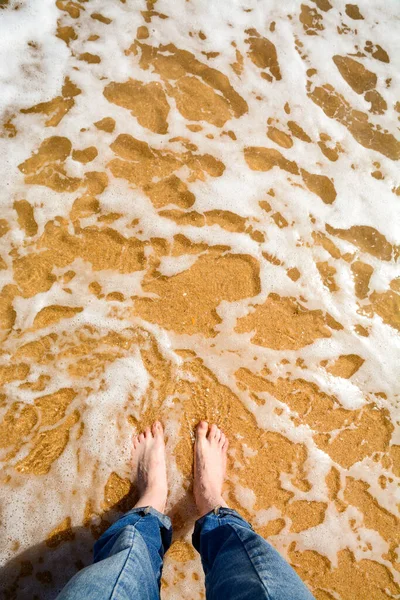 Bare feet on sandy beach. Ocean water touches feet. Waves cover bare feet of game