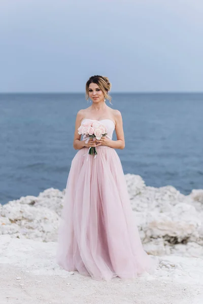 Πορτρέτο μιας νεαρής όμορφης νύφης με ένα μπουκέτο τριαντάφυλλα κοντά στη θάλασσα. — Φωτογραφία Αρχείου