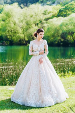 Gelin, yeşil çimlerin ve gölün arkasında beyaz bir gelinlik giyiyor..