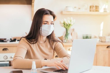 Maskeli kız evde, mutfakta dizüstü bilgisayarla çalışıyor. Uzaktan kumanda, koronovirüs yüzünden izolasyon. Merkez ofis. Bağımsız ve uzak çalışma