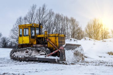 Yeni inşaat alanında sarı kazıcı, parlak güneş ve arka planda güzel mavi gökyüzü. Kışın sokakta sarı buldozer, kar yağışı ve kar yamaçları.