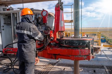Kırmızı üniformalı, miğferli ve gözlüklü matkapçı. Petrol kuyusuna indirmek için matkap borularını vidalamak ve delmeye devam etmek için hidrolik anahtar kullanıyor. Çalışan insan kavramı.