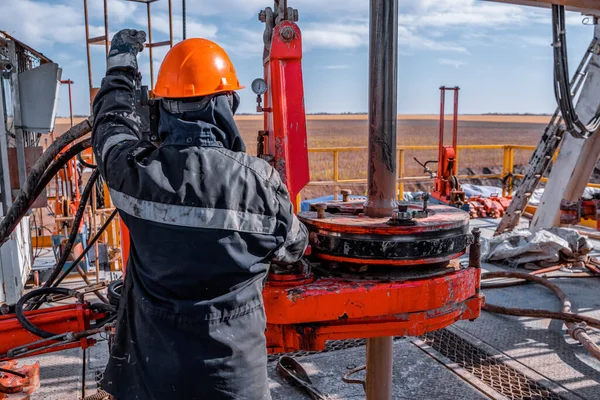 Taladro de trabajo en uniforme rojo, en casco y gafas. Utiliza una llave hidráulica para atornillar tubos de perforación para bajarlos a un pozo de petróleo y continuar perforándolo. Concepto de trabajador. —  Fotos de Stock