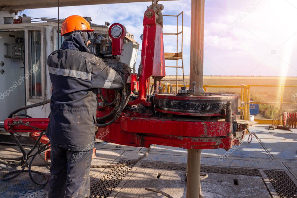 Work driller in red uniform, in helmet and goggles. He uses a hydraulic wrench to screw drill pipes to lower them into an oil well and continue drilling it. The concept of a working person.