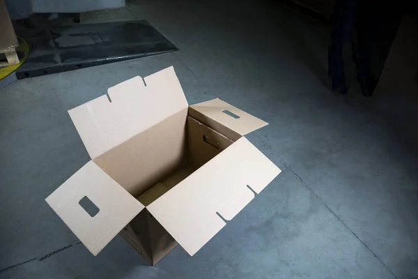 A big, open, empty cardboard box standing on the stony floor in a warehouse.