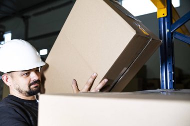 Elinde karton bir kutu tutan kasklı bir depo çalışanı. Kutuları yığının üzerine koymak. Fabrikada güvenli bir iş..