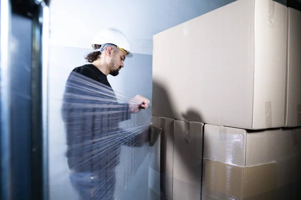 Karton kutuları esnek bir folyoya sarmak. Fabrikadaki depoda kutu paketleyen bir adam..