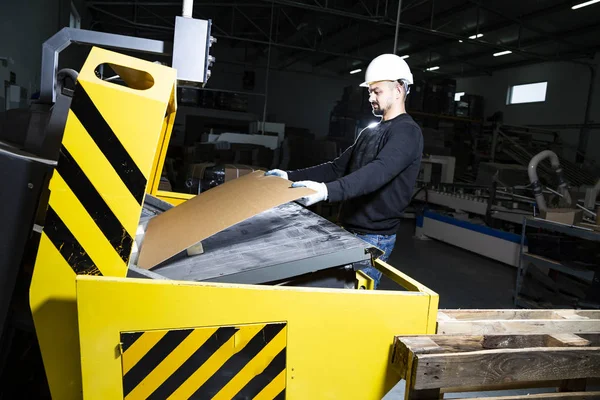 Cardboard boxes production hall. Man in hard hat putting carton into a punching machine. Paper die cutting machine.