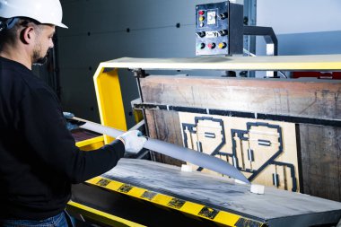 Sert şapkalı bir işçi bir karton kutuyu yumruk makinesine yerleştiriyor. Karton kutu fabrikası. Kağıt kesme makinesi