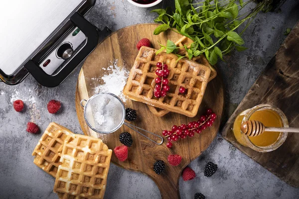 Freshly baked waffles, fruits, powdered sugar, honey and a professional waffle iron, top view. — Stock Photo, Image