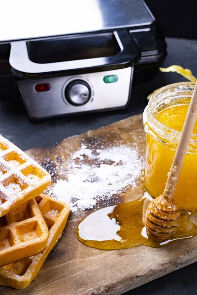 Freshly baked waffles with powdered sugar and honey. Waffle iron in a background. — Stock Photo, Image