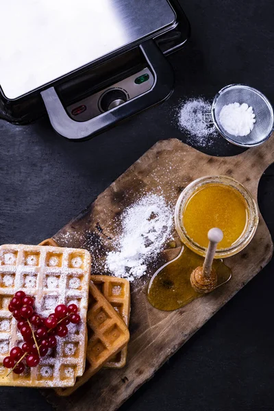 Golden, baked waffles, fruits, powdered sugar, honey and a professional waffle iron, top view. — Stock Photo, Image