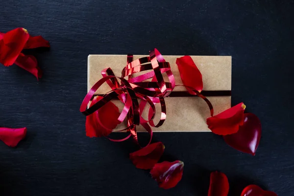 Valentine's Day, love. Red roses petals, heart and gift with a red ribbon on a black background — Stockfoto