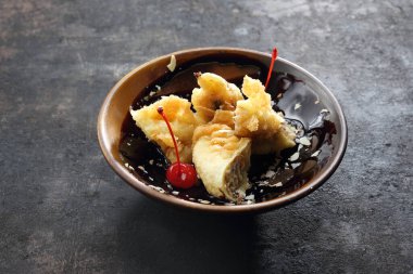 Çikolata soslu tempura içinde kızarmış muz, badem gevreği ve maraska vişneli küçük, minimal tarzda kahverengi bir kâse. Siyah taşlı arka planda doğulu bir tatlı..