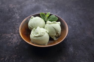 Üç kepçe yeşil çay, şam fıstığı, nane yaprağıyla süslenmiş oryantal dondurma küçük, minimal tarzda, kahverengi bir kâse. Koyu taşlı arka planda doğulu bir tatlı. 