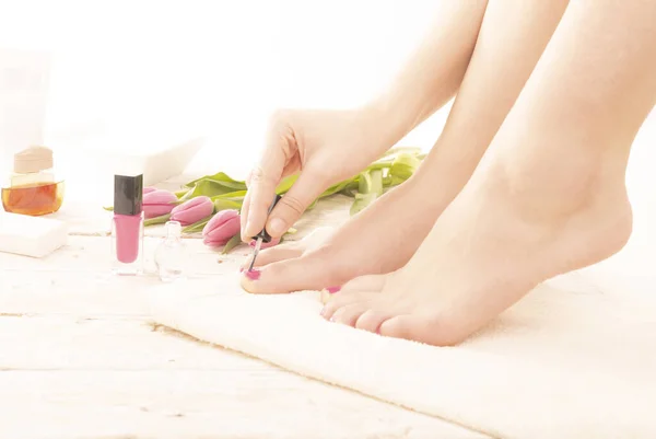 SPA pedicure. A young woman painting fingernails with pink nail polish. Body care, foot relaxing in cozy place with wood floor decorated with tulips. Wellness, day SPA, beauty salon. Women\'s Day