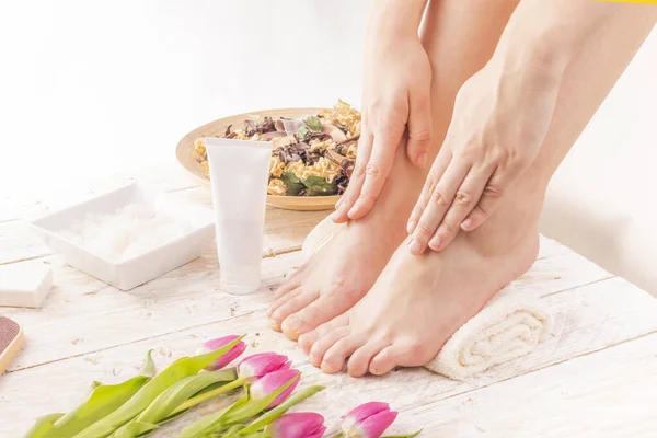 A young woman putting cream on her feet placed on a towel. Body care, SPA pedicure, foot massage in a cozy place decorated with pink tulips. Wellness and day SPA, beauty salon. Women\'s Day.