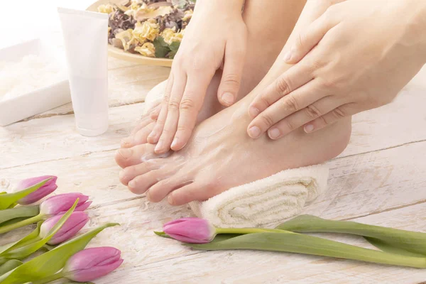 A young woman putting cream on her feet placed on a towel. Body care, SPA pedicure, foot massage in a cozy place decorated with pink tulips. Wellness and day SPA, beauty salon. Women\'s Day.