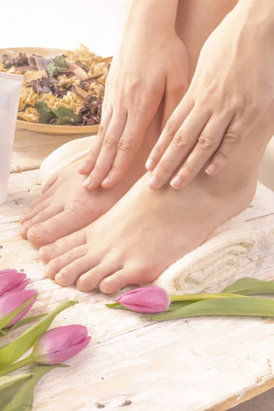 A young woman putting cream on her feet placed on a towel. Body care, SPA pedicure, foot massage in a cozy place decorated with pink tulips. Wellness and day SPA, beauty salon. Women's Day.