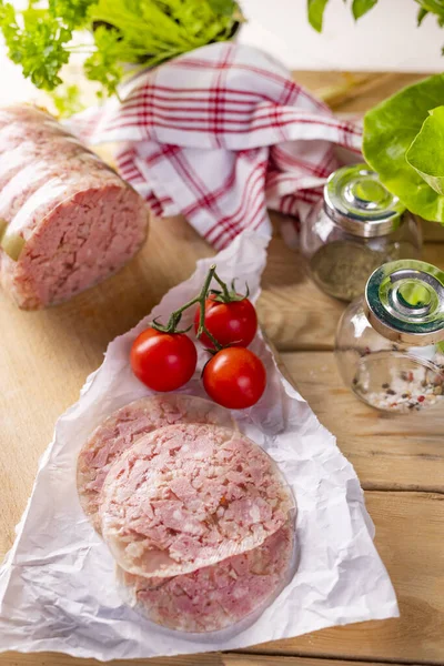 Headcheese cut into slices. Pork cold cuts. Sliced brawn on a paper, on wood worktop. Rustic composition of headcheese, fresh herbs, tomatoes, spices, kitchen towel and wood boards. Pork meat delicacy