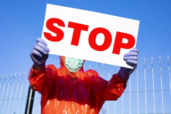 STOP. A man in a protective suit, wearing gloves, glasses, a surgical mask, holding information board and stopping drivers and passers-by on a street on a sunny day. Stop corona, SARS, virus pandemic.