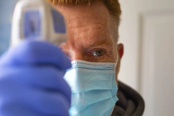 Redhead man in nitrile protective gloves and single use surgical mask, measuring body temperature with an infrared forehead thermometer. Fighting coronavirus pandemic from China.
