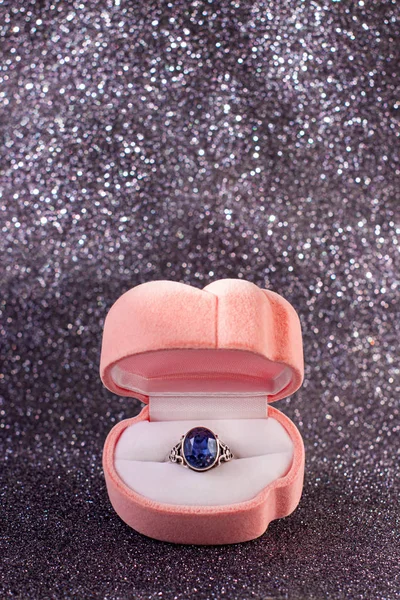 Pink velvet box with antique silver ring with blue shiny gem on black glitter background, front view. St. Valentines Day