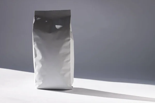 Бланширующая белая сумка из фольги на монохромном фоне в минимальном стиле с естественной светлой тенью. Упаковка для продуктов питания или напитков — стоковое фото