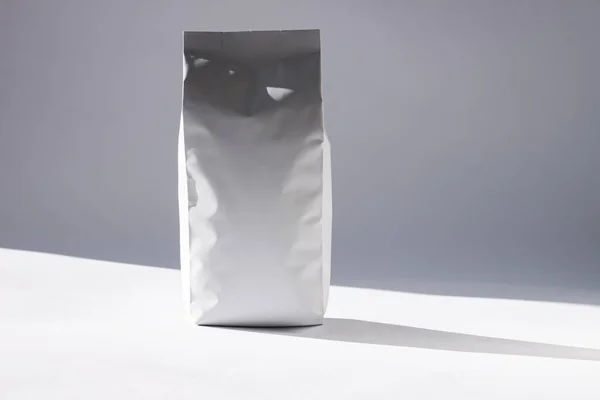 Бланширующая белая сумка из фольги на монохромном фоне в минимальном стиле с естественной светлой тенью. Упаковка для продуктов питания или напитков — стоковое фото