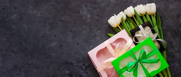 White tulip flowers and colorful gift boxes with ribbon bows on black background flat lay. Website banner header.Place for text 8March Happy Womens Mothers Day.Flower Bouquet greeting card. Copy space