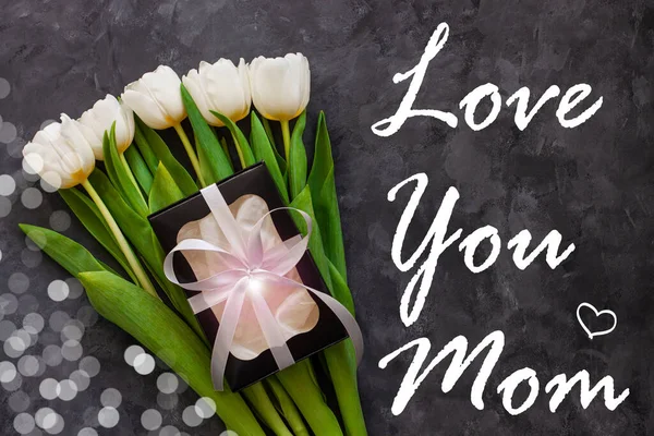 Love You Mom greeting card. White tulip flowers and black gift box on dark background flat lay.Place for text 8 March Womens Mothers Day.Flower Bouquet silver ribbon.Copy space website banner top view