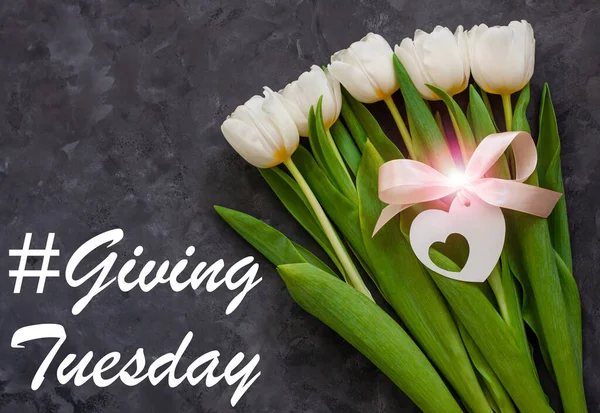 Giving Tuesday card message sign. Charity Donations concept. White tulip flowers and heart card tag with pink ribbon bow on dark background flat lay. Flower Bouquet with bokeh lights top view.