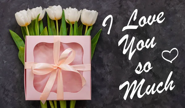 I Love You So Much card sign text. White tulip flowers and pink gift box with ribbon bow on dark background flat lay. Female flower Bouquet greeting card. Copy space website banner top view.