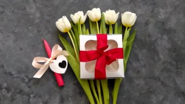 Beyaz lale çiçekleri, kırmızı kurdeleli gümüş hediye kutusu ve koyu arka plandaki mum 4K üst manzaralı. Doğum günün kutlu olsun 8 Mart Kadınlar Anneler Günü konsepti. Bahar çiçeği buketi hediye, kadın blog