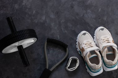 Spor ekipmanları düz konum, beyaz spor ayakkabı, spor ayakkabı, spor saati, fitness göğüs genişletici, karın bölgesi mide eğitimi için karın çarkları gri beton arka plan. Sağlıklı yaşam tarzını üst bakış açısıyla kavra