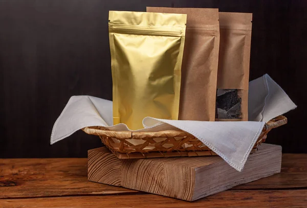 Paper and foil pouch bags with coffee beans and tea leaves in wicker basket with white textile on wooden background. Packaging for foods and goods mockup. Weight products packs with clasps and windows