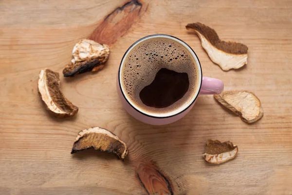 Mushroom coffee chaga superfood. Dried mushrooms and fresh brewed pink cup of hot drink with foam on wooden background top view. Healthy organic energizing adaptogen, endurance boosting food trends.