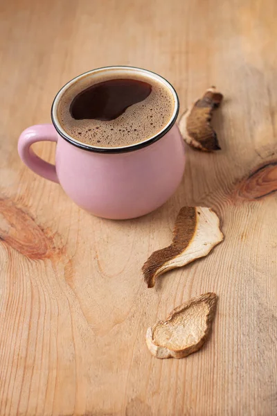 Mushroom coffee chaga superfood. Dried mushrooms and fresh brewed pink cup of hot drink with foam on wooden background vertical photo.Healthy organic energizing adaptogen,endurance boosting food trend