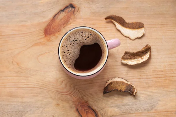 Mushroom coffee chaga superfood. Dried mushrooms and fresh brewed pink cup of hot drink with foam on wooden background top view. Healthy organic energizing adaptogen, endurance boosting food trends.