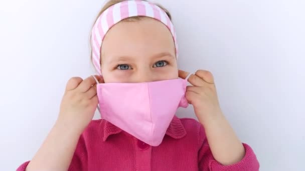 頭皮の小さな女の子は コロナウイルスCovid 19パンデミック白の背景に対して保護ピンクのファブリックマスクを身に着けています 子供の触れる顔 手作りの縫製保護マスク — ストック動画