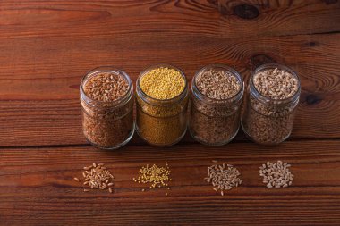 Tahta arka planda cam kavanozlardaki tahıl taneleri. Farklı Groats koleksiyonları. Arpa, yulaf, darı ve buğday. Yüksek lif içerikli kepekli gıdalar düz yerde yatıyordu. Sağlıklı beslenme malzemeleri.