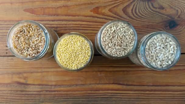 木製の背景4Kビデオ上のガラス瓶の穀物 オート麦 小麦を表示するさまざまな大麦のコレクション 全粒食品高繊維含有量フラット層 健康的な食事成分 — ストック動画