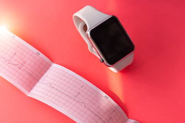 Beyaz spor silikon bantlı akıllı saat, pembe mercan arka planlı ECG diyagram kağıdı. Kardiyogram tıbbi muayene. Kalp hastalığını önler, mitral kapakçık kaymasını önler. Kalp atışlarını analiz eder..