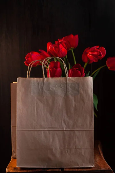 Kraft paper bags with blooming red tulips bouquet on black background. Dark moody low key minimalism style flowers and food delivery packaging banner copy space mockup. Donations help recycling boxes.