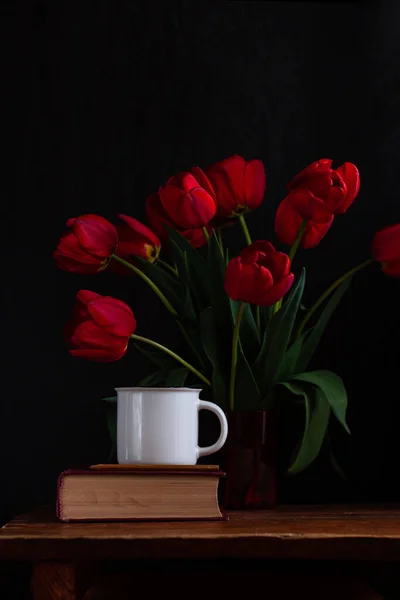 木製の鉛筆で古い本の上に白いコーヒーカップ 黒の背景に驚くべき開花赤いチューリップ花束 レトロな静物 良い朝のバナー — ストック写真