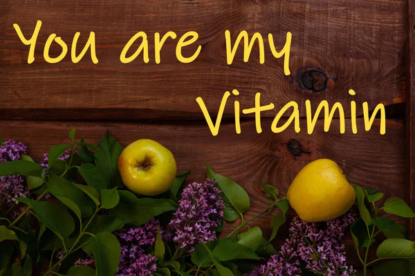 You are my Vitamin text sign. Bright yellow apples, purple lilac flowers with fresh green leaves on wooden background flat lay. Colorful spring summer juicy fruit backdrop. Cotrasting creative banner.