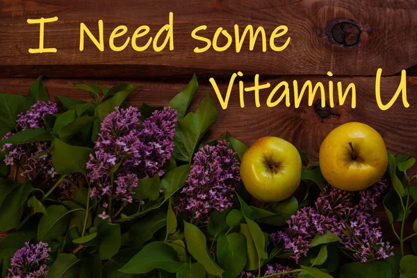 I need some Vitamin U text. Bright yellow apples, purple lilac flowers with fresh green leaves on wooden background flat lay. Colorful Love spring summer fruit backdrop. Cotrasting creative banner.