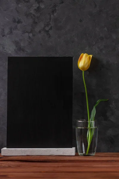 Blank chalkboard mockup and yellow tulip in a water glass on dark wooden background. Blackboard with beautiful spring flower wallpaper copy space. Minimalism style. Text sign greeting card.