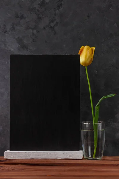 Blank chalkboard mockup and yellow tulip in a water glass on dark wooden background. Blackboard with beautiful spring flower wallpaper copy space. Minimalism style. Text sign greeting card.